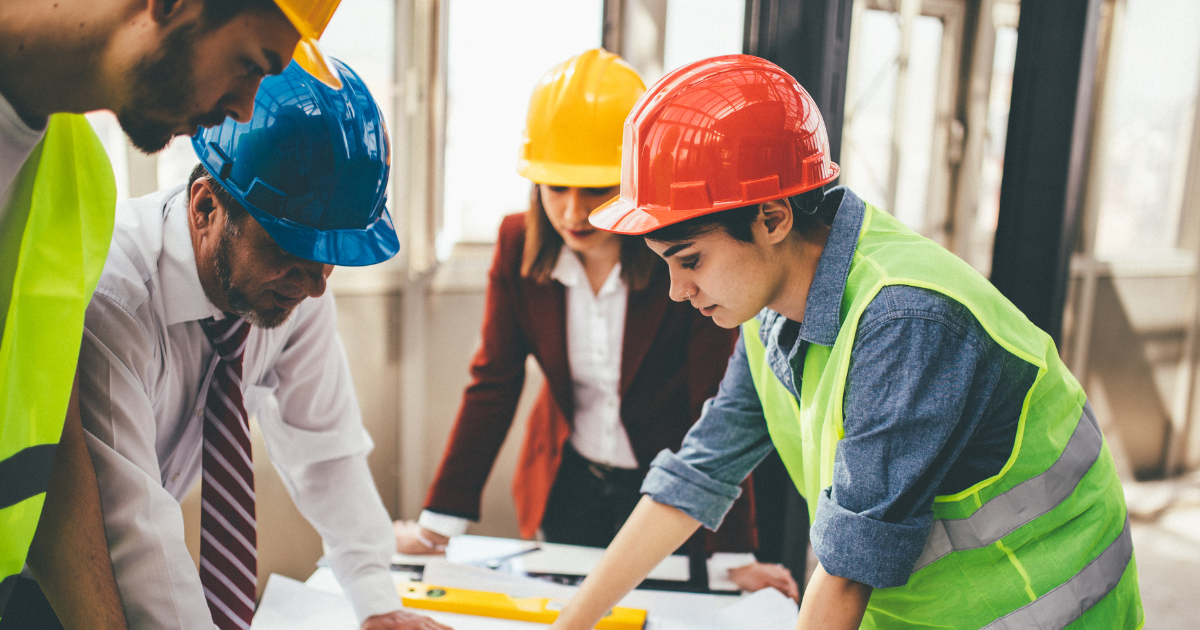 FWR 3 Website (1200 x 630 px) The image shows four construction workers gathered around a table, examining plans or documents. They are indoors with large windows in the background, letting in ample natural light. All four individuals are wearing safety helmets and appear focused on the task at hand. The person on the left, wearing a white hard hat and safety vest, is looking down at the plans. The person in the middle wears a yellow hard hat, with their attention directed at the documents. A person to the right is wearing a red hard hat and a bright green safety vest, closely examining the documents with a forward lean. The fourth person in the background, wearing a blue hard hat, is also engaged in the discussion.