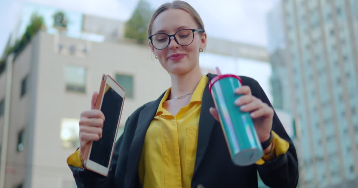 A person with glasses holding a smartphone and a colorful travel mug, standing outdoors with buildings in the background.