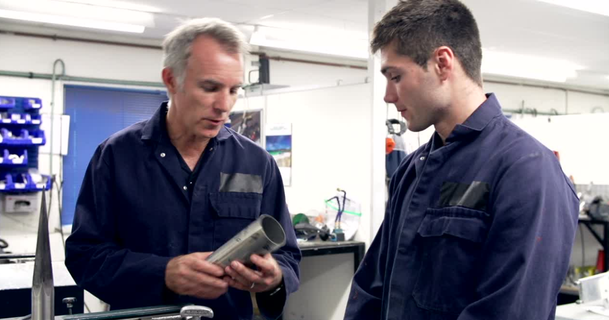The image depicts two men in a workshop or industrial setting. Both are wearing dark blue coveralls. The man on the left, with gray hair, is holding a cylindrical metal object and appears to be explaining something to the man on the right, who has brown hair. They are engaged in conversation, and the background features workshop equipment, including shelves filled with blue storage bins, tools on a workbench, and some machinery.