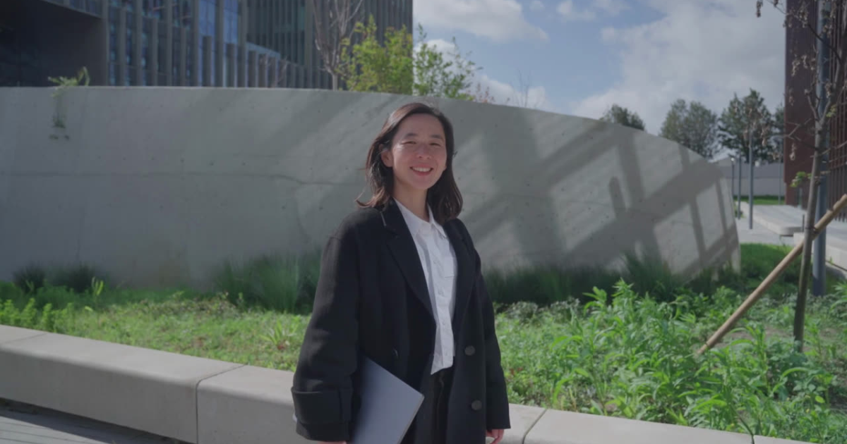 The image shows a person standing outdoors in a sunny setting. They have shoulder-length dark hair and are wearing a black coat over a white shirt, while holding a closed laptop at their side. The background features a concrete wall with shadows cast across it, surrounded by lush green plants. A building with vertical design elements is visible in the left background, and the sky is partly cloudy with blue patches.