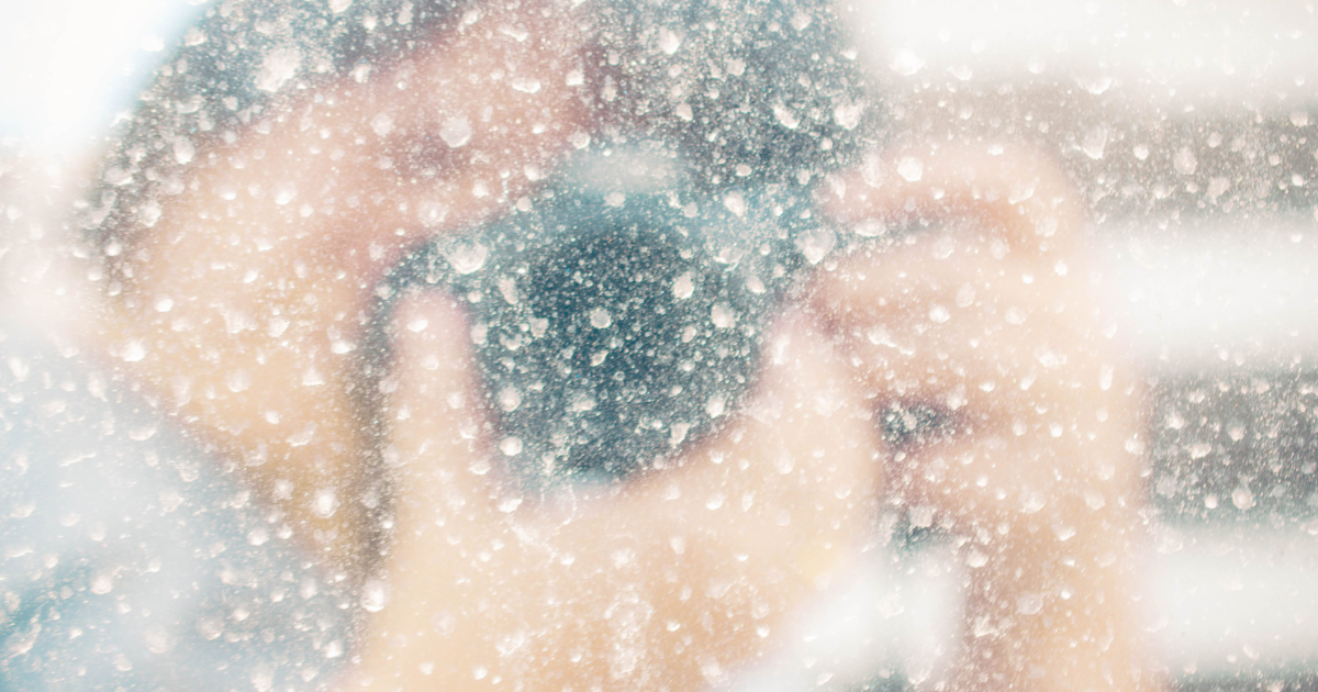 Blurred view of a person with a camera behind a water-splattered glass.