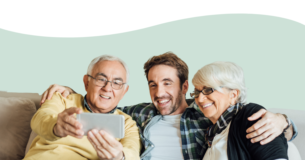 A decorative photo of a young man on a sofa between his older parents, they are smiling at a cell phone.